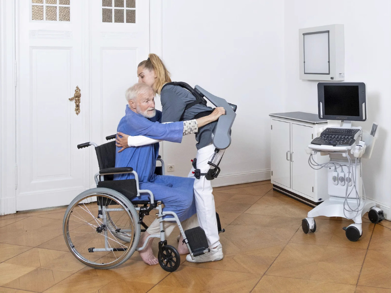Person assisting an elderly person in a wheelchair with a robotic exoskeleton.